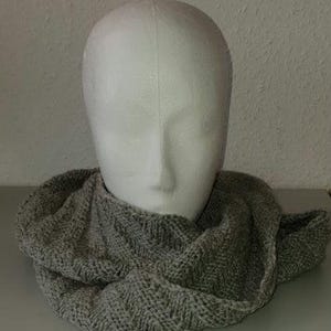Op de afbeelding: Een grijze gebreide infinity sjaal op een witte mannequin hoofd. De sjaal heeft een textuur, geweven patroon en is gemaakt van zacht, neutraal getint garen. De sjaal is om de nek van de mannequin gewikkeld.
