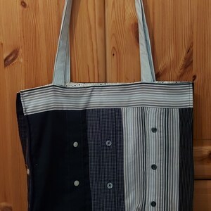 Könnte beinhalten: Eine Tragetasche aus wiederverwendetem Stoff. Die Tasche ist aus verschiedenen gemusterten Hemden gefertigt, mit einem blau-weiß gestreiften Stoff oben und unten. Die Tasche hat zwei lange Henkel und einen flachen Boden.
