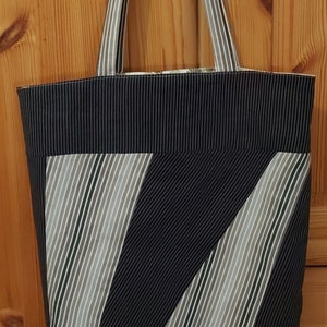 Könnte beinhalten: Eine schwarz-weiß gestreifte Tragetasche mit einem geometrischen Muster aus diagonalen Streifen. Die Tasche hat zwei lange Henkel.