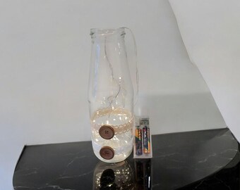 May include: A clear glass bottle with a cork top and a string of lights inside. The bottle is decorated with two wooden buttons and a piece of lace. The bottle is sitting on a black surface.