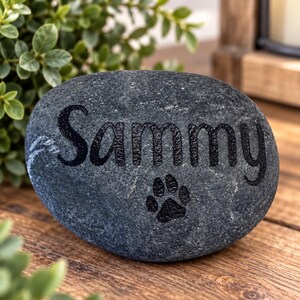 May include: A smooth, dark grey river stone featuring the name Sammy engraved in a black, sans-serif font. Below the name, a small black paw print icon is also engraved into the surface of the stone, creating a clear and simple memorial marker.