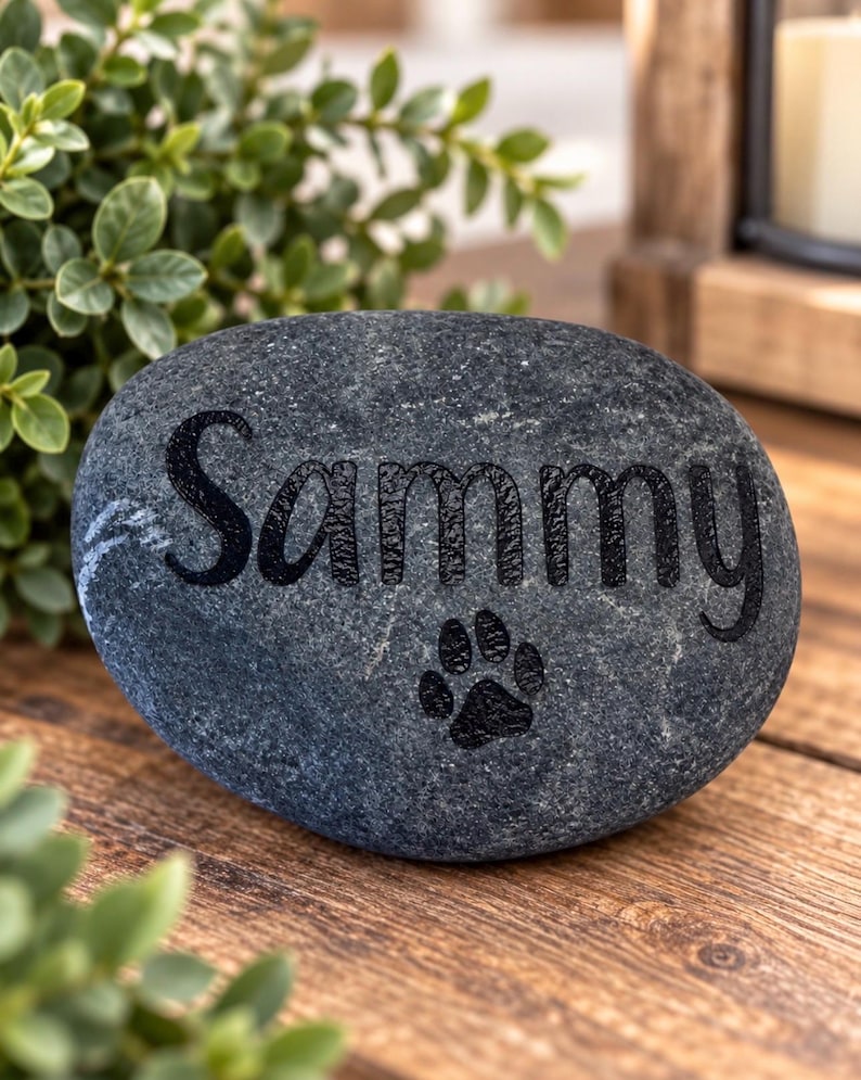 May include: A smooth, dark grey river stone featuring the name Sammy engraved in a black, sans-serif font. Below the name, a small black paw print icon is also engraved into the surface of the stone, creating a clear and simple memorial marker.