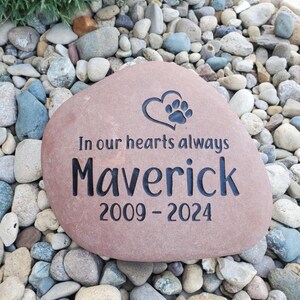 May include: A smooth, tan-coloured memorial stone featuring an engraved heart and paw print symbol. The text reads In our hearts always, the name Maverick, and the years 2009-2024. The lettering and graphic are deeply etched in a dark, contrasting colour.
