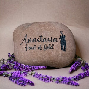 May include: A brown stone with the name Anastasia and the phrase "Heart of Gold" engraved on it. A black silhouette of an elephant is also engraved on the stone. The stone is surrounded by purple flowers.