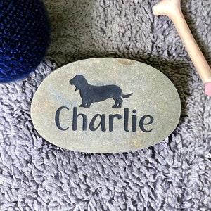 May include: A gray oval-shaped stone with a black silhouette of a dachshund and the name "Charlie" engraved on it.