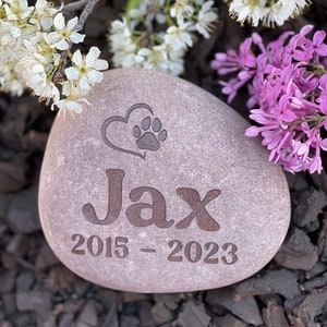 May include: A gray memorial stone with the name "Jax" and the years "2015-2023" engraved on it. A heart with a paw print is above the name. The stone is surrounded by white and purple flowers.