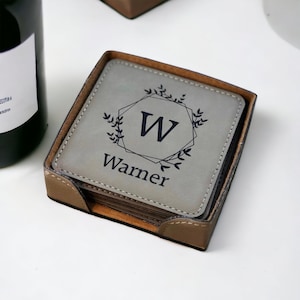 May include: A set of square grey coasters with stitched edges, housed in a brown holder. The top coaster features a black printed design with a geometric frame, leafy accents, the letter W, and the name Warner centred in a serif font.