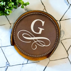 May include: A brown leather coaster with a stitched edge and a white monogram of the letter "G" with a decorative swirl design.