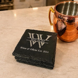 May include: A stack of four square black slate coasters with rough, natural edges. The top coaster is engraved with a large letter W, the name WILSON, and the text Brian & Olivia Est. 2014. A shiny copper mug with a gold handle sits nearby.