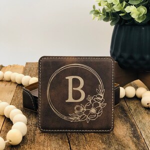 May include: A square dark brown leather coaster featuring a laser-engraved design of the letter B centred within a circular frame, accented by a floral motif on the bottom right corner, with visible white stitching along the edges.