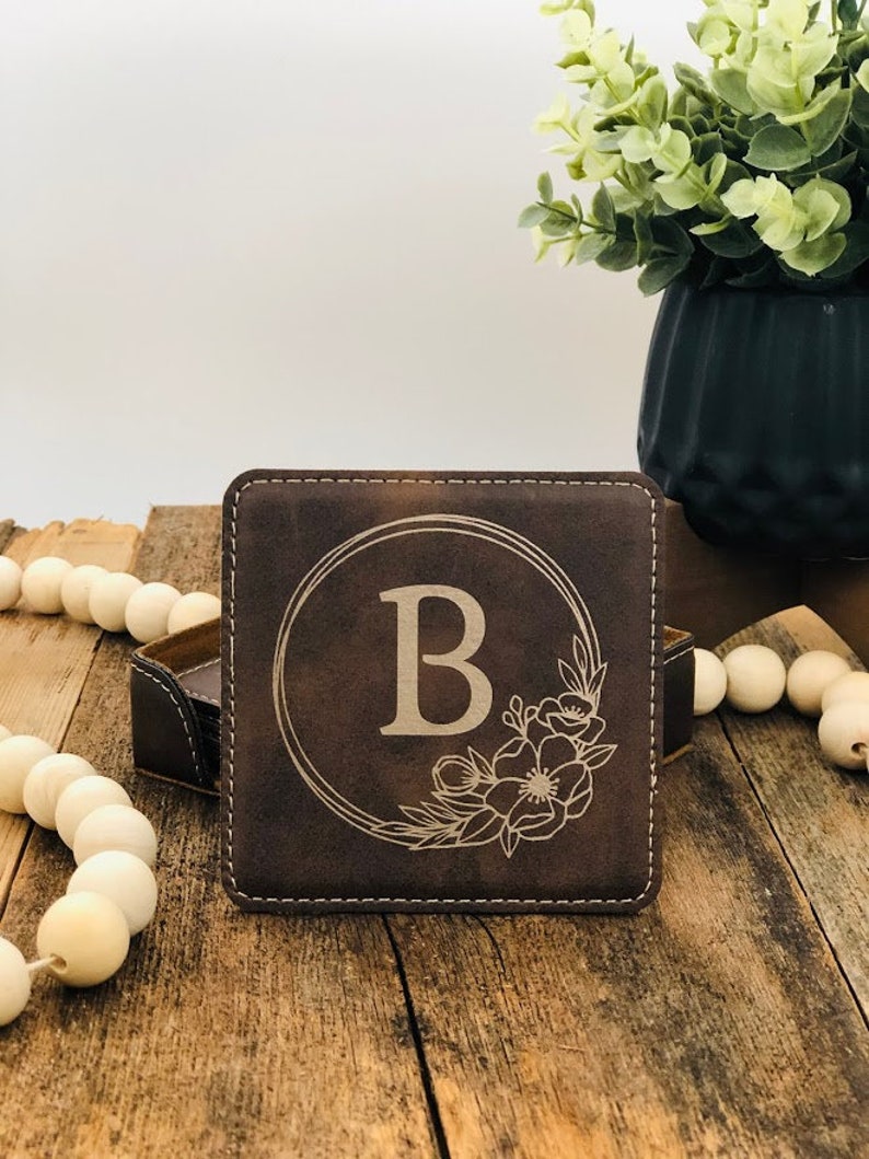 May include: A square dark brown leather coaster featuring a laser-engraved design of the letter B centred within a circular frame, accented by a floral motif on the bottom right corner, with visible white stitching along the edges.
