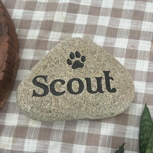 May include: A smooth, light grey river stone featuring the name Scout engraved in black lettering. Above the name, a black paw print icon is centred. The stone has a natural, rounded triangular shape with a textured, speckled surface finish.