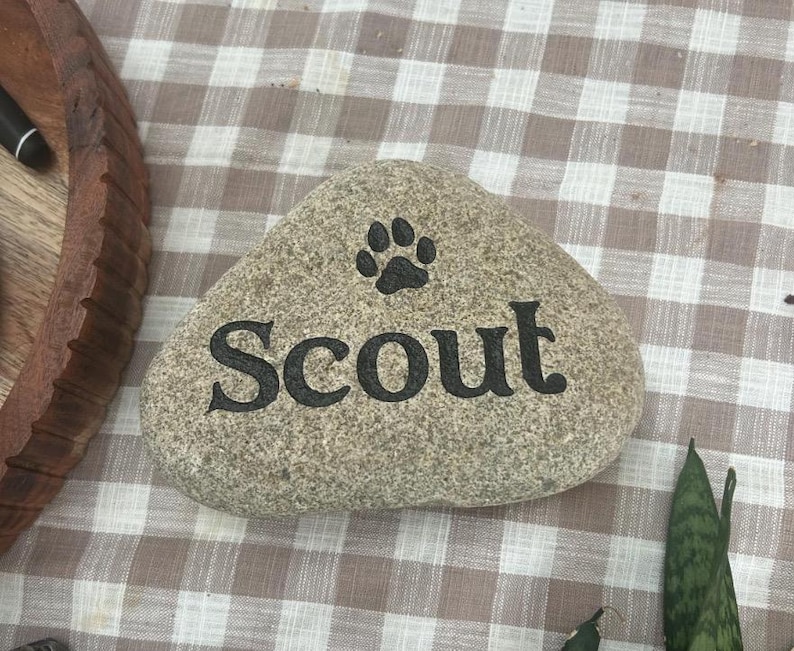 May include: A smooth, light grey river stone featuring the name Scout engraved in black lettering. Above the name, a black paw print icon is centred. The stone has a natural, rounded triangular shape with a textured, speckled surface finish.