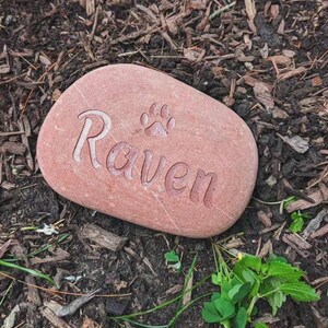 May include: A smooth, oval-shaped reddish-brown stone featuring the engraved name Raven in a cursive font, accompanied by a small paw print icon etched into the surface. The stone has a natural texture and is positioned on a dark soil surface.