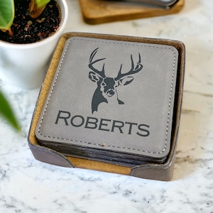 May include: A stack of square grey coasters with rounded corners housed in a dark brown holder. Each coaster features a black laser-engraved graphic of a deer head silhouette above the capitalised text ROBERTS in a clean sans-serif font.