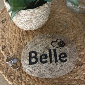 May include: A smooth, speckled grey river stone engraved with the name Belle in black lettering, accompanied by a black graphic of a heart and a paw print. Next to the stone is a silver-toned metal ring featuring a small, round, light-coloured stone.