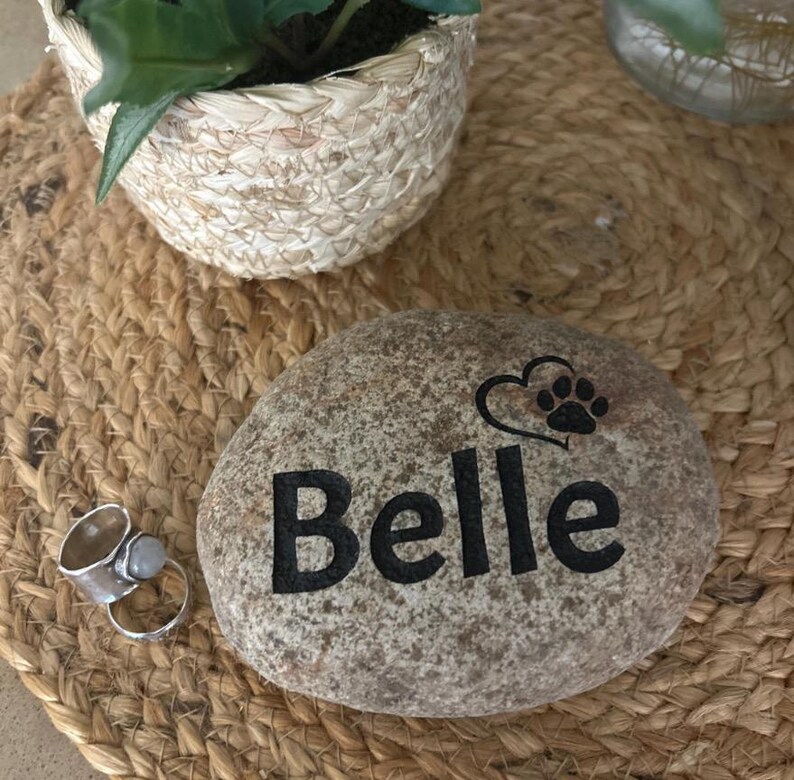 May include: A smooth, speckled grey river stone engraved with the name Belle in black lettering, accompanied by a black graphic of a heart and a paw print. Next to the stone is a silver-toned metal ring featuring a small, round, light-coloured stone.