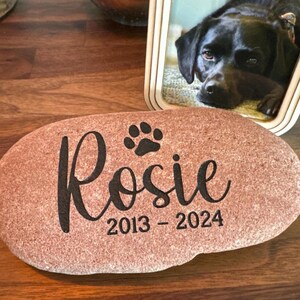 May include: A smooth, oval-shaped reddish-brown memorial stone featuring the engraved name Rosie in a cursive script, a small paw print icon above the name, and the years 2013 - 2024 engraved in a clean, sans-serif font below the name.