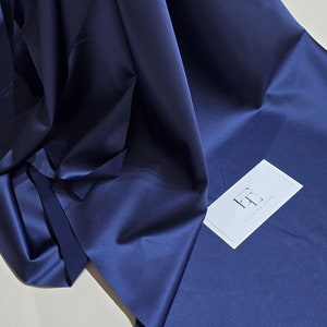 Könnte beinhalten: Dunkelblaues Satin-Gewebe, drapiert und gefaltet, wodurch Schatten und Licht entstehen. Das Material hat einen seidigen Glanz. Eine weiße Visitenkarte mit dem Text "LACE TO LOVE" liegt auf dem Stoff.