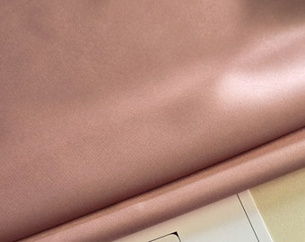 Puede incluir: Primer plano de una tela de satén rosa pálido doblada sobre una tarjeta blanca con el texto "Lace to Love" y un monograma negro de las letras "LT" en un cuadrado.