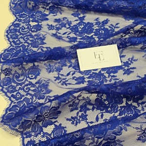 May include: A close-up shot of a royal blue lace fabric featuring a detailed floral design. The fabric has a scalloped edge and is layered. A white card with the text "LACE TO LOVE" is present.