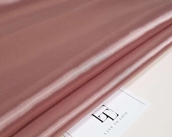 Puede incluir: Primer plano de una tela de satén rosa empolvado doblada. La tela tiene una superficie lisa y brillante con pliegues y arrugas visibles. Una tarjeta blanca con el texto "LACE TO LOVE" es parcialmente visible.