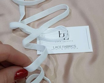 May include: A length of white elastic ribbon coiled in loops. A white rectangular business card is present with the black logo and text: LACE TO LOVE, LACE FABRICS, Retail & Wholesale. The ribbon has a smooth, matte texture and consistent width.