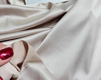 Puede incluir: Primer plano de una tela fluida de color beige suave, con pliegues y sombras suaves. La tela tiene un brillo lujoso. Una pequeña tarjeta con el texto "LACE TO LOVE" es visible en la parte superior de la imagen.