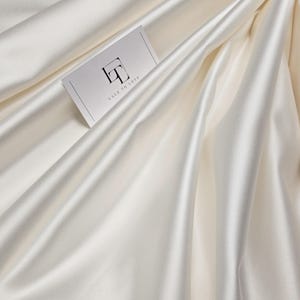 May include: Ivory-colored satin fabric with soft folds and a lustrous sheen. A business card with the text "LACE TO LOVE" and a stylized logo is placed on the fabric. The fabric appears smooth and luxurious.