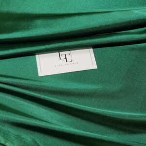 May include: A close-up of a deep green fabric with a subtle sheen. The fabric is folded and draped, showcasing its texture. A white label with the text "Lace To Love" is visible in the center of the image.