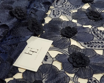 May include: A navy blue lace fabric featuring a repeating floral pattern with three-dimensional embroidered flower centers. A white rectangular tag with the black text "LT LACE TO LOVE" is placed on top of the fabric, which has a sheer mesh base.