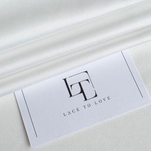 Könnte beinhalten: Eine weiße Visitenkarte mit dem Text "LACE TO LOVE" und einem stilisierten schwarzen "LT"-Logo. Die Karte liegt auf einem weißen, seidigen Stoff mit sanften Falten. Die Gesamtästhetik ist sauber und elegant.