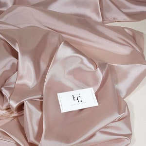 Könnte beinhalten: Nahaufnahme eines blushrosa Satinstoffs mit weichen Falten und einem glänzenden Glanz. Eine weiße Visitenkarte mit dem Text "LACE TO LOVE" liegt auf dem Stoff. Der Stoff wird wahrscheinlich für Kleidung oder Heimdekoration verwendet.