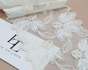 Op de afbeelding: Een brede strook gebroken witte kanten stof met een ingewikkeld geborduurd bloemen- en bladpatroon met geschulpte randen. Een witte rechthoekige kaart is gedeeltelijk eronder zichtbaar met de zwarte tekst LT LACE TO LOVE.