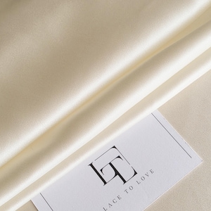 May include: Ivory-colored satin fabric with soft folds and a subtle sheen. A white card with the text "LACE TO LOVE" and a stylized logo is partially visible. The fabric's texture suggests a luxurious textile.