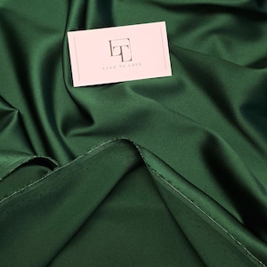 Könnte beinhalten: Smaragdgrüner Satin-Stoff mit weichen Falten und dezentem Glanz. Eine hellrosa Visitenkarte mit dem Text "LACE TO LOVE" liegt auf dem Stoff. Das Material eignet sich für Bekleidung oder Heimtextilien.