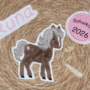 Könnte beinhalten: Ein braunes Pferde-Applikation mit grauer Mähne und Schweif, einem rosa Herz und einem weißen Rand. Das Bild enthält auch eine weiße Applikation mit dem Namen "Runa" in Rosa und ein rosa-weißes Etikett mit "Schulkind 2026".