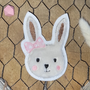 Könnte beinhalten: Ein weißes Hasengesicht aus Filz mit einer rosa Schleife, rosigen Wangen und einer schwarzen Nase und Augen. Der Hase hat glitzernde braune Innenohren. Der Hase befindet sich auf einem Holzgrund mit Drahtgeflecht.