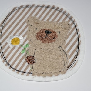 Könnte beinhalten: Ein gestickter Aufnäher mit einem Teddybären, der eine Blume hält. Der Bär ist braun und weiß, der Hintergrund ist ein braunes und weißes Streifenmuster.