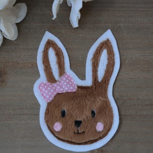 Könnte beinhalten: Ein braunes Kaninchen mit weißen Ohren und einer rosa Schleife mit weißen Punkten. Das Kaninchen hat einen weißen Umriss und befindet sich auf einem braunen Holzgrund.