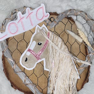 Könnte beinhalten: Wanddekoration mit einem Pferdeapplikation, einem rosa Glitzerhalfter und dem Namen "Lotta" in rosa Schrift. Das Design befindet sich auf einer Holzscheibe, umrahmt von einem Drahtherz.