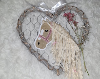 May include: A beige horse head with a pink bridle is attached to a heart-shaped wire wreath. The wreath is made of twigs and has a white ribbon hanging from the top.