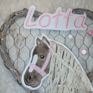 Könnte beinhalten: Ein brauner Filz-Pferdekopf mit einem rosafarbenen und weißen Zaumzeug und einer langen weißen Mähne. Der Pferdekopf ist auf einem weißen Filz-Hintergrund befestigt. Der Name "Lotta" ist in rosa Stickerei auf einem weißen Filz-Hintergrund geschrieben.