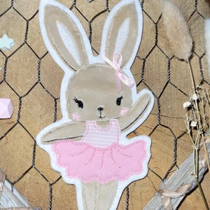 May include: A brown felt bunny wearing a pink tutu and a pink bow. The bunny is embroidered with white details and has a striped bodice.