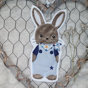 Könnte beinhalten: Ein braunes Kaninchen mit weißen Ohren und einer blau-weiß gestreiften Latzhose. Das Kaninchen trägt ein blaues Hemd mit einem weißen und gelben Sternenmuster-Halstuch.