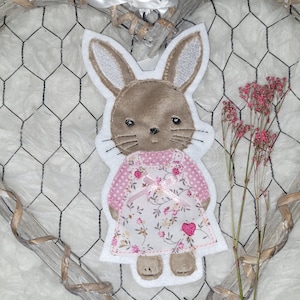 Könnte beinhalten: Ein braunes Filzkaninchen, das ein rosa-weißes Blumenkleid mit einer rosa Schleife trägt. Das Kaninchen steht auf einem weißen Hintergrund mit einem Drahtgitterherz dahinter.