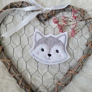 Könnte beinhalten: Ein graues und weißes gesticktes Wolfskopf-Applik mit rosa Wangen. Das Applik ist an einem herzförmigen Holzrahmen mit einem weißen Band befestigt.