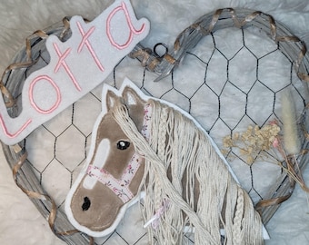 May include: A decorative heart-shaped wall hanging with a wire frame and a plush horse head. The horse head is tan with a white mane and a floral patterned bridle. The name "Lotta" is embroidered in pink on a white banner.