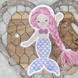 Könnte beinhalten: Eine rosa Filz-Meerjungfrauenpuppe mit geflochtenem rosa Haar, einem blau-rosa Meerjungfrauen-Schwanz und einem rosa Muschel-BH. Die Puppe befindet sich auf einer Holzoberfläche mit einem Drahtgitter-Hintergrund.