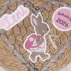 Könnte beinhalten: Ein herzförmiger Wandschmuck mit einem braunen Filzhasen in einem rosa Blumenkleid mit Rucksack, einem weißen Filzschild mit dem Namen Leni in rosa Schrift und einem runden weißen Filzanhänger mit rosa Herzen und der Aufschrift Schulkind 2026.